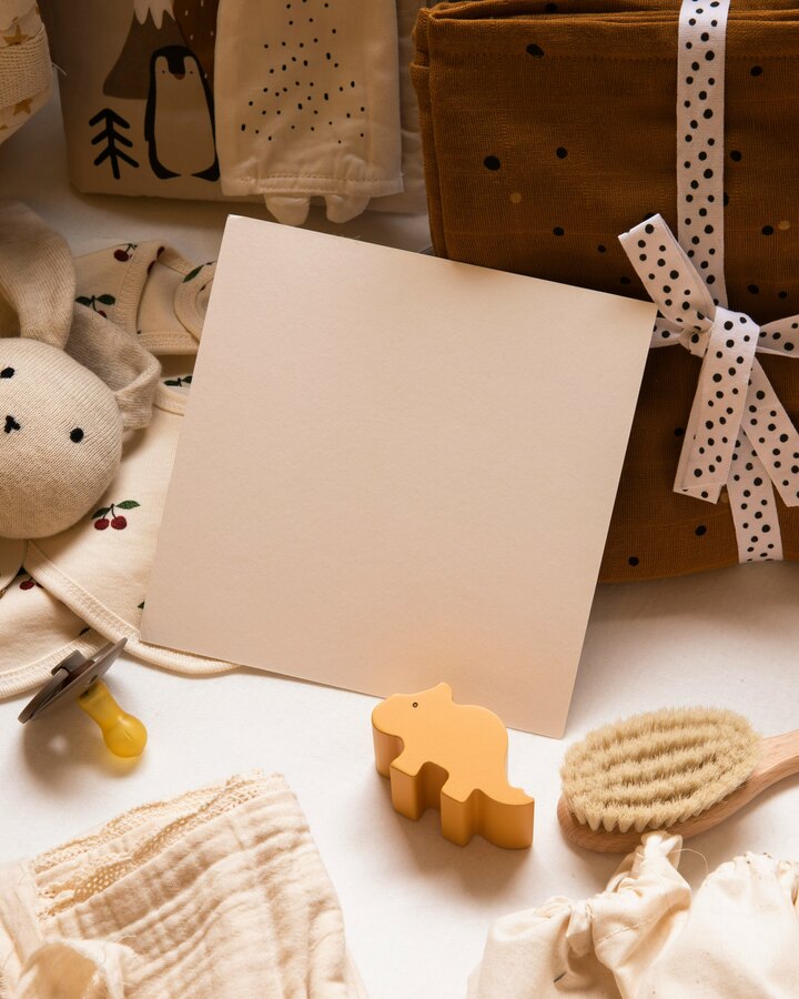 A neutral-toned flat-lay of baby items — a cream knit bunny rattle, a cherry-print onesie, a wooden brush, muslin wraps tied with polka-dot ribbon, and a blank cream card.