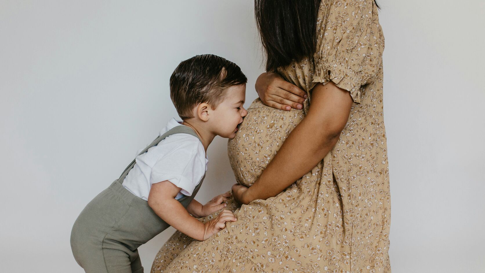 A toddler in a white t-shirt and grey suspender shorts leans in to kiss his mother's pregnant belly, her hands resting on the bump in a cream floral dress.