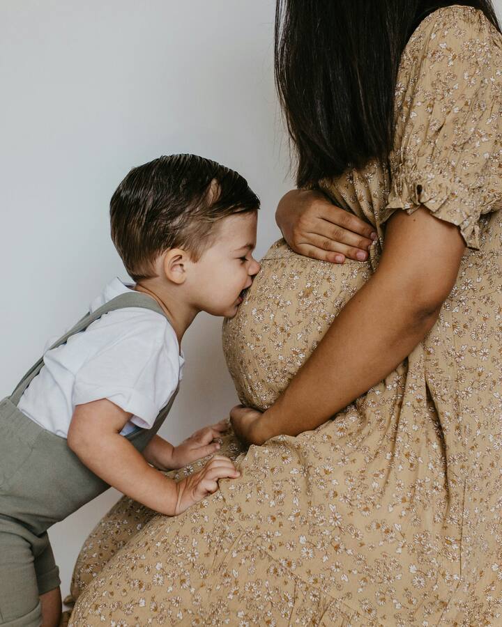 A toddler in a white t-shirt and grey suspender shorts leans in to kiss his mother&rsquo;s pregnant belly, her hands resting on the bump in a cream floral dress.