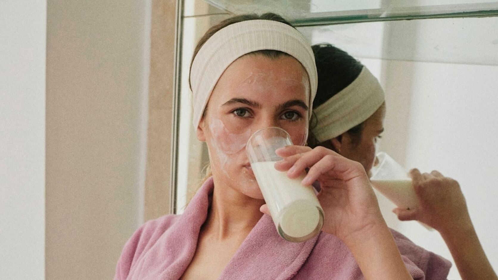A woman in a headband and pink robe with a cream face mask on, sipping a glass of milk in front of a bathroom mirror.