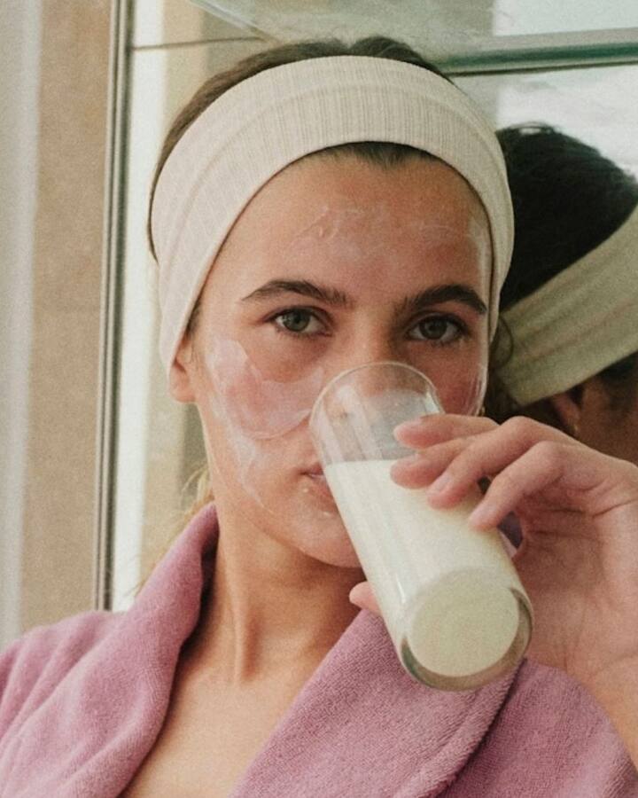 A woman in a headband and pink robe with a cream face mask on, sipping a glass of milk in front of a bathroom mirror.