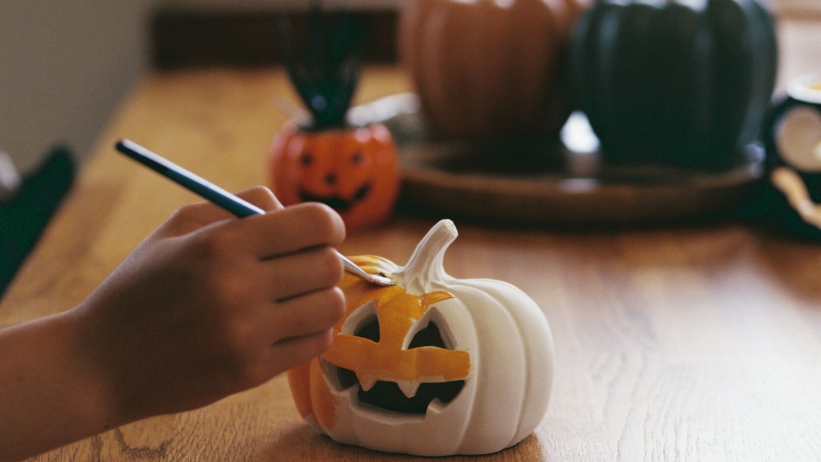 A child&rsquo;s hand paints the carved grin of a small white jack-o&rsquo;-lantern orange with a fine brush on a wooden table, with an orange, a rust and a dark green pumpkin blurred behind.