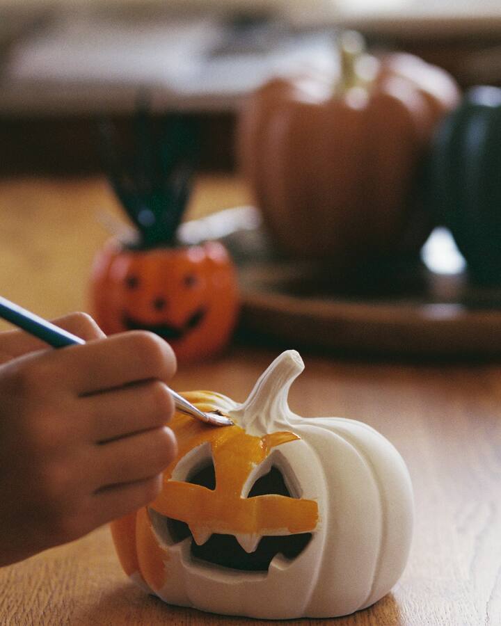 A child&rsquo;s hand paints the carved grin of a small white jack-o&rsquo;-lantern orange with a fine brush on a wooden table, with an orange, a rust and a dark green pumpkin blurred behind.