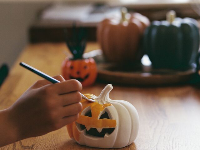 A child&rsquo;s hand paints the carved grin of a small white jack-o&rsquo;-lantern orange with a fine brush on a wooden table, with an orange, a rust and a dark green pumpkin blurred behind.