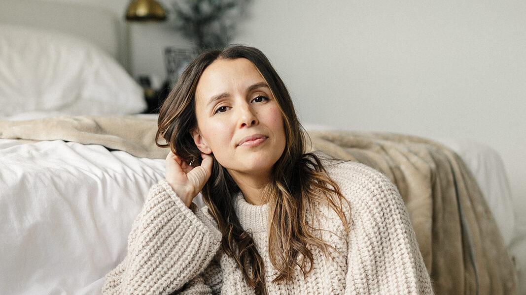 Rachael Shepard-Ohta sits on the floor beside her bed in an oatmeal knit sweater and wide-leg jeans, one hand in her hair, looking at the camera.