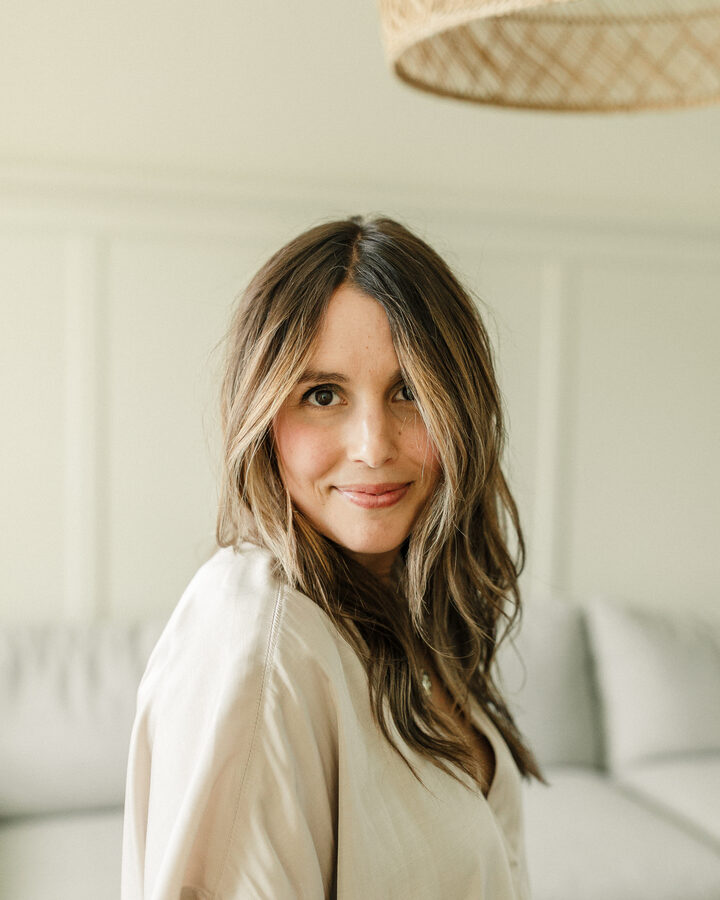 Rachael Shepard-Ohta in a soft cream top, photographed in a bright living room, looking over her shoulder at the camera.