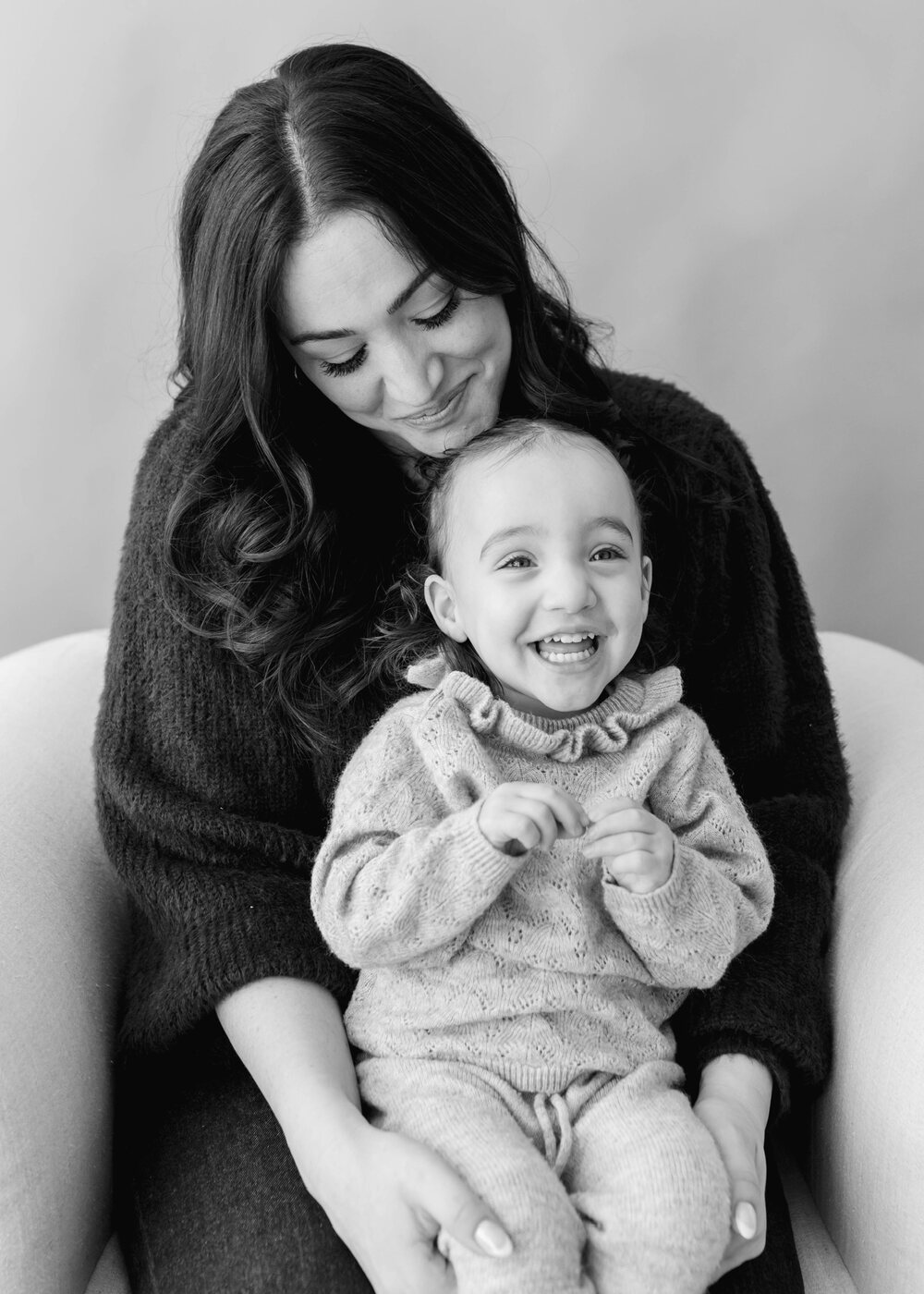 A black-and-white studio portrait of Rachel Abugov looking down at her laughing toddler daughter Brooklyn on her lap.