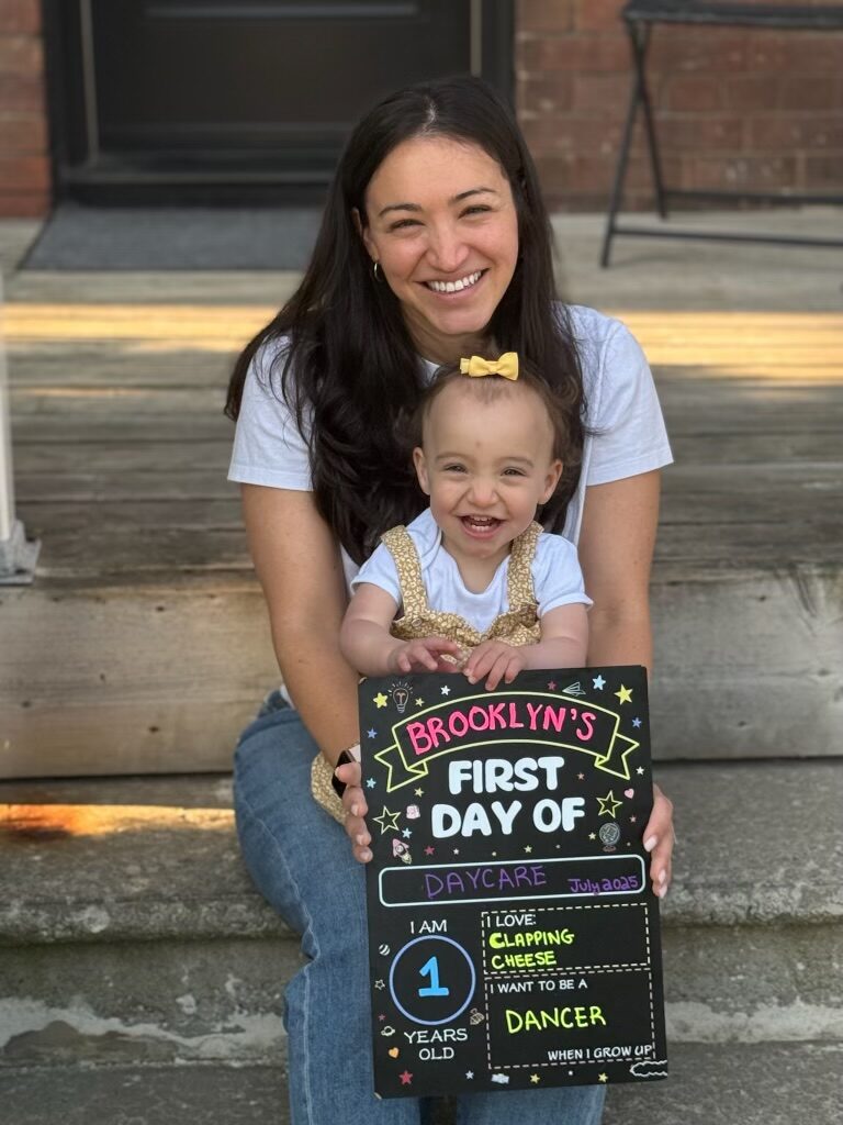 Rachel sits on her front steps with a thriving one-year-old Brooklyn, holding a chalkboard sign for Brooklyn's first day of daycare.
