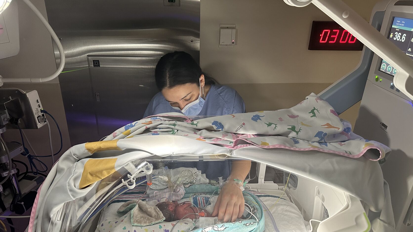 Rachel Abugov leans over an incubator in the NICU, her hand resting beside her tiny newborn daughter, Brooklyn.