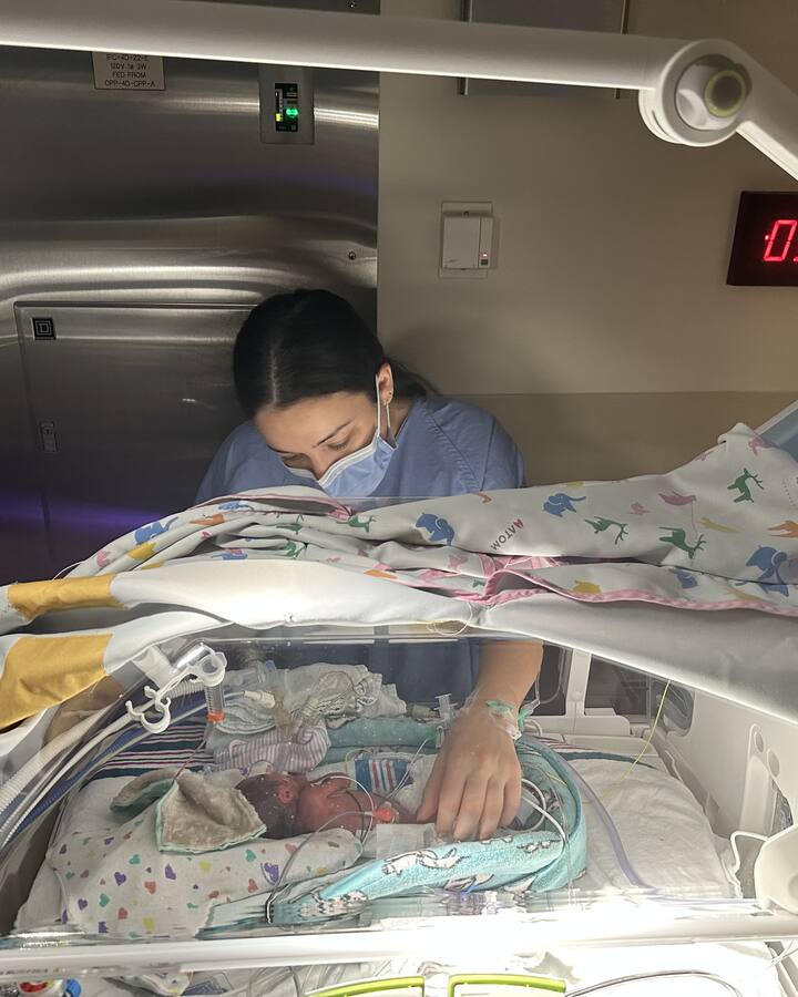 Rachel Abugov leans over an incubator in the NICU beside her tiny newborn daughter, Brooklyn.