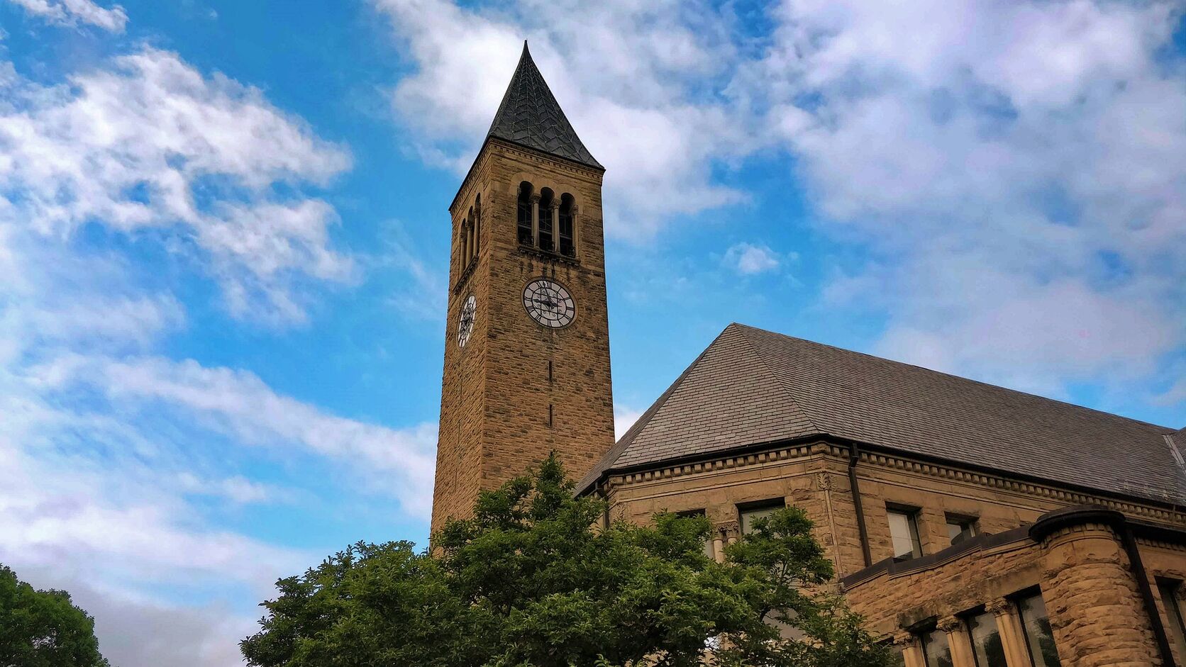 Cornell University&rsquo;s McGraw Tower, a stone clock tower with a dark spire, rises above leafy trees and a sandstone building against a bright blue sky with drifting clouds.
