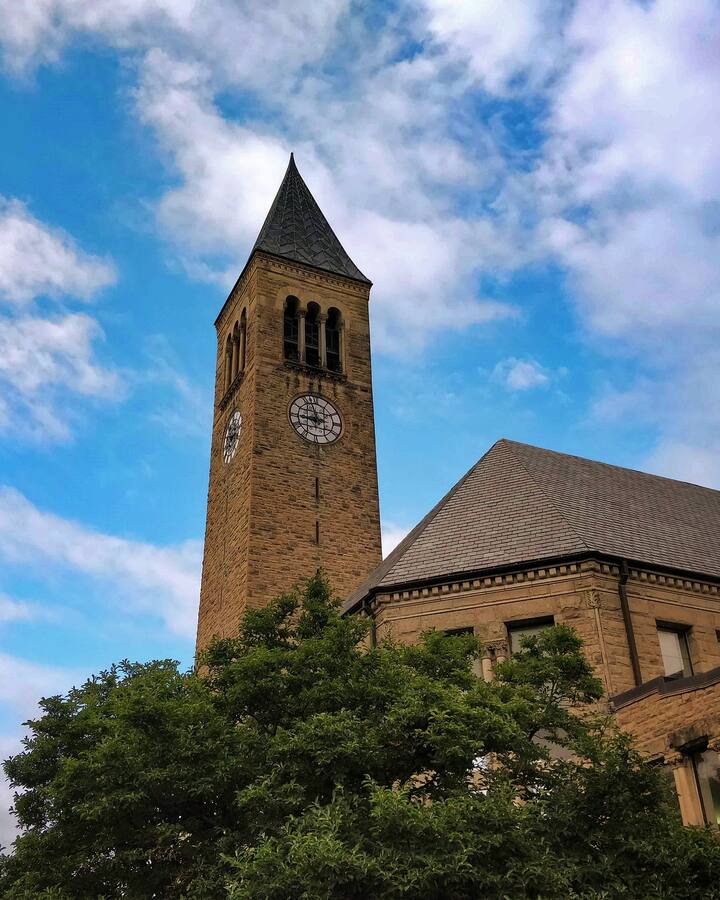 Cornell University&rsquo;s McGraw Tower, a stone clock tower with a dark spire, rises above leafy trees and a sandstone building against a bright blue sky with drifting clouds.
