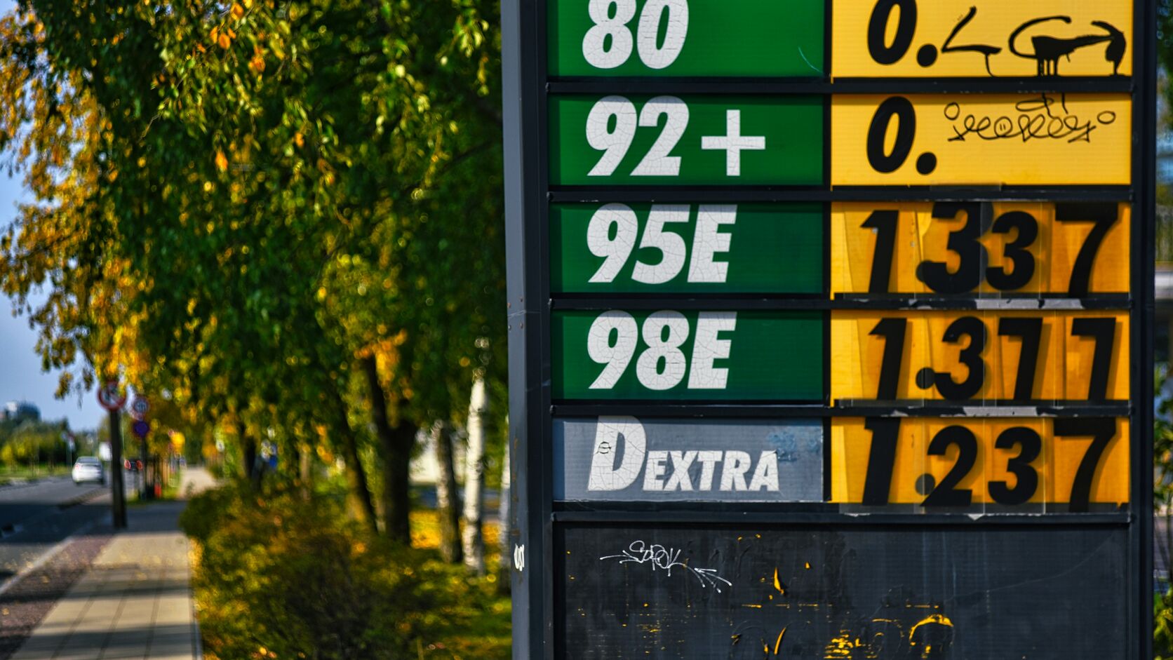 A roadside fuel-price sign in green and yellow lists steadily rising prices beside a tree-lined autumn sidewalk.