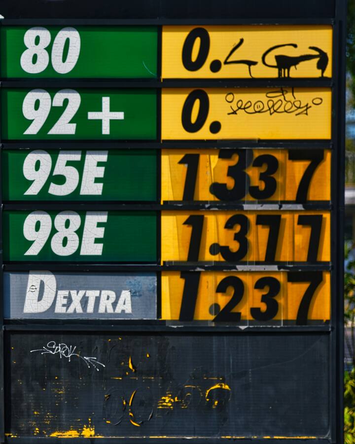 A roadside fuel-price sign in green and yellow lists steadily rising prices beside a tree-lined autumn sidewalk.