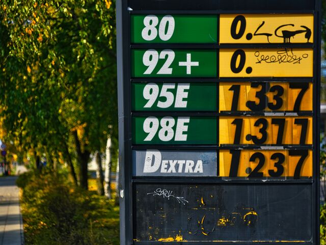 A roadside fuel-price sign in green and yellow lists steadily rising prices beside a tree-lined autumn sidewalk.