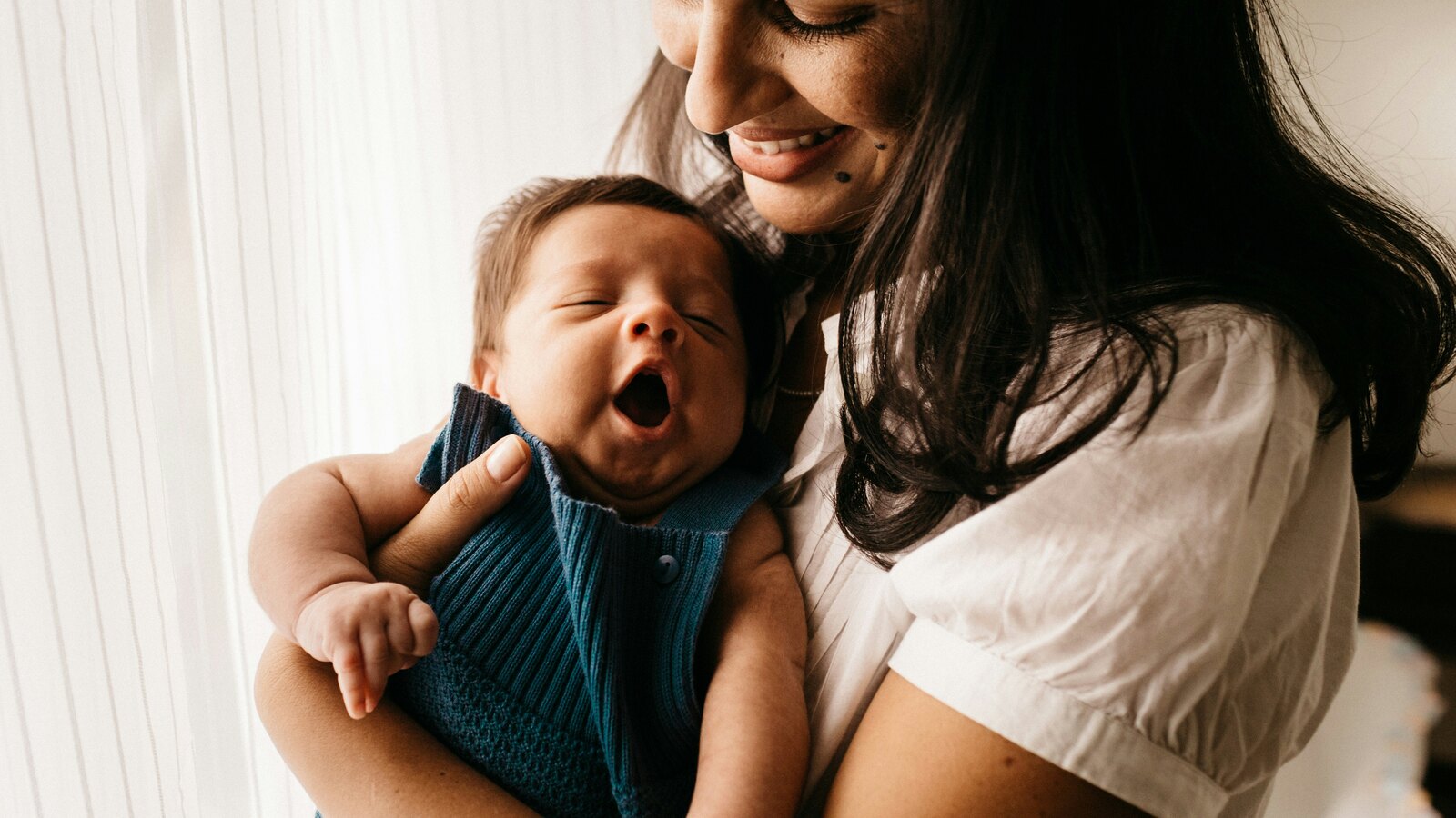 A smiling mother in a white shirt holds her yawning newborn, dressed in blue knit, close to her chest by a bright window.