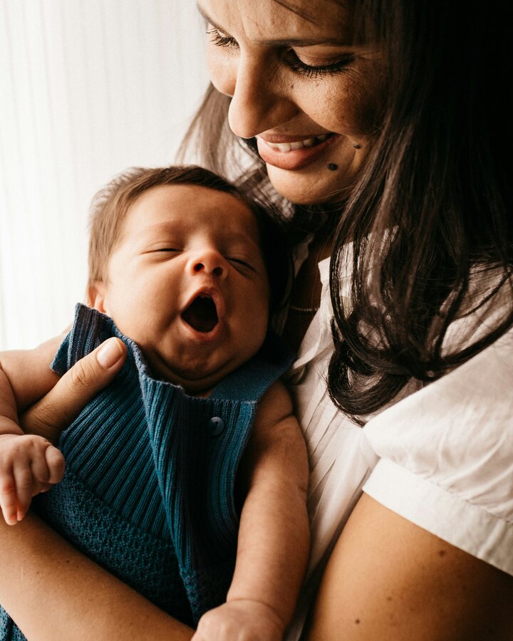 A smiling mother in a white shirt holds her yawning newborn, dressed in blue knit, close to her chest by a bright window.