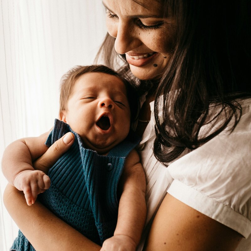 A smiling mother in a white shirt holds her yawning newborn, dressed in blue knit, close to her chest by a bright window.