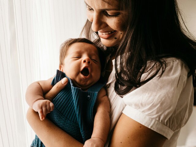 A smiling mother in a white shirt holds her yawning newborn, dressed in blue knit, close to her chest by a bright window.