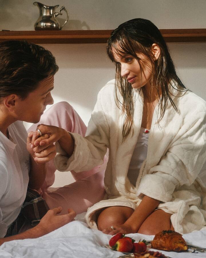 A couple sit close together on a bed with a breakfast tray, as he gently holds her hand to his lips and she smiles softly toward him.