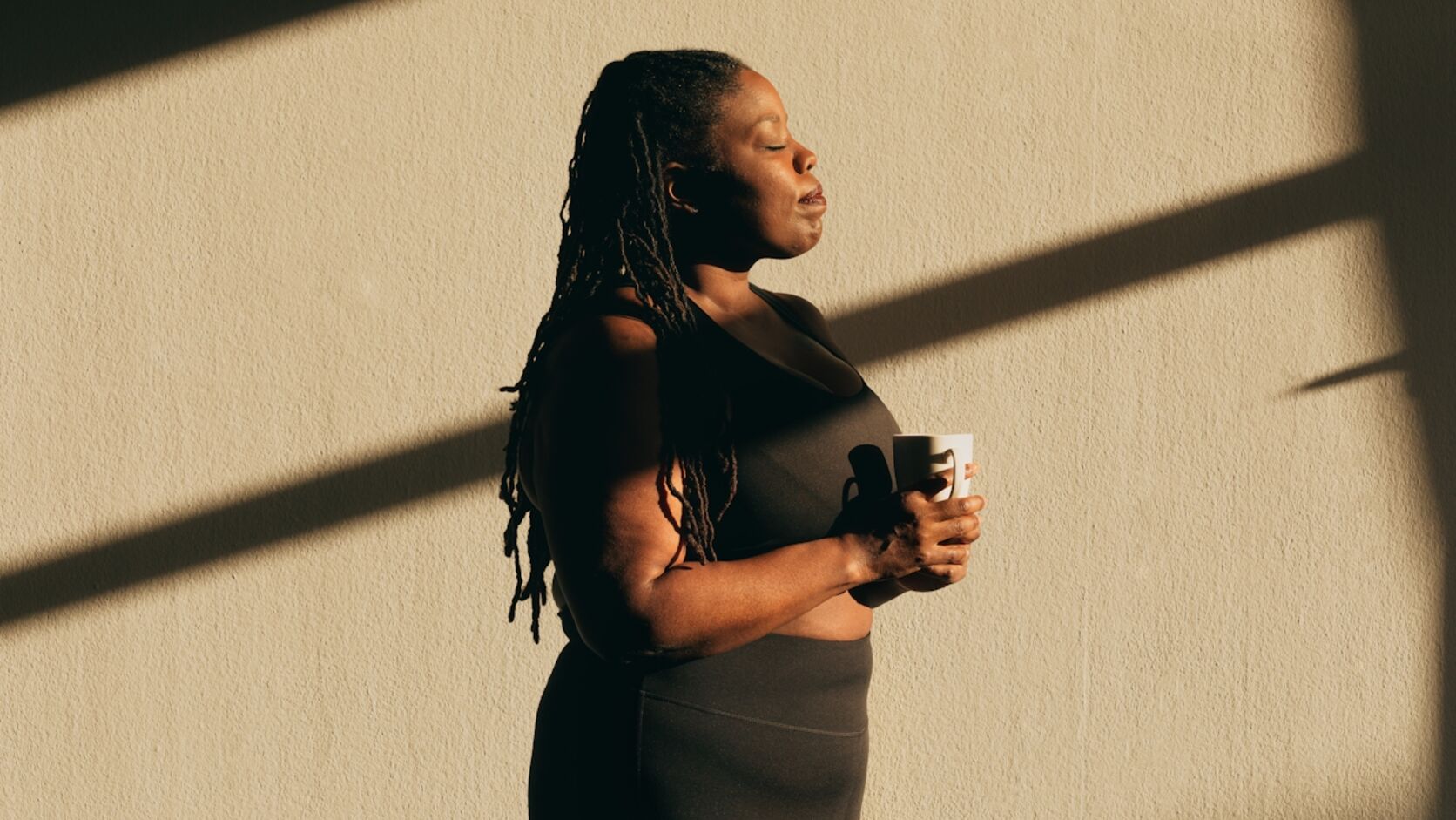 A woman in athletic wear stands against a sunlit wall with her eyes closed, holding a mug in both hands in a quiet moment to herself.