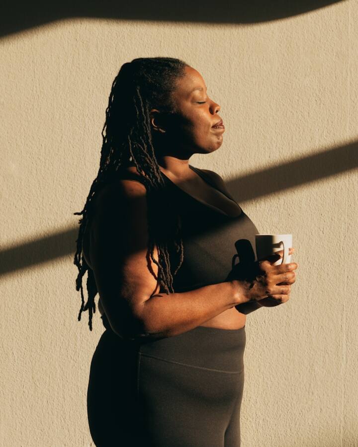 A woman in athletic wear stands against a sunlit wall with her eyes closed, holding a mug in both hands in a quiet moment to herself.