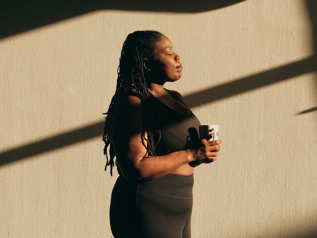 A woman in athletic wear stands against a sunlit wall with her eyes closed, holding a mug in both hands in a quiet moment to herself.