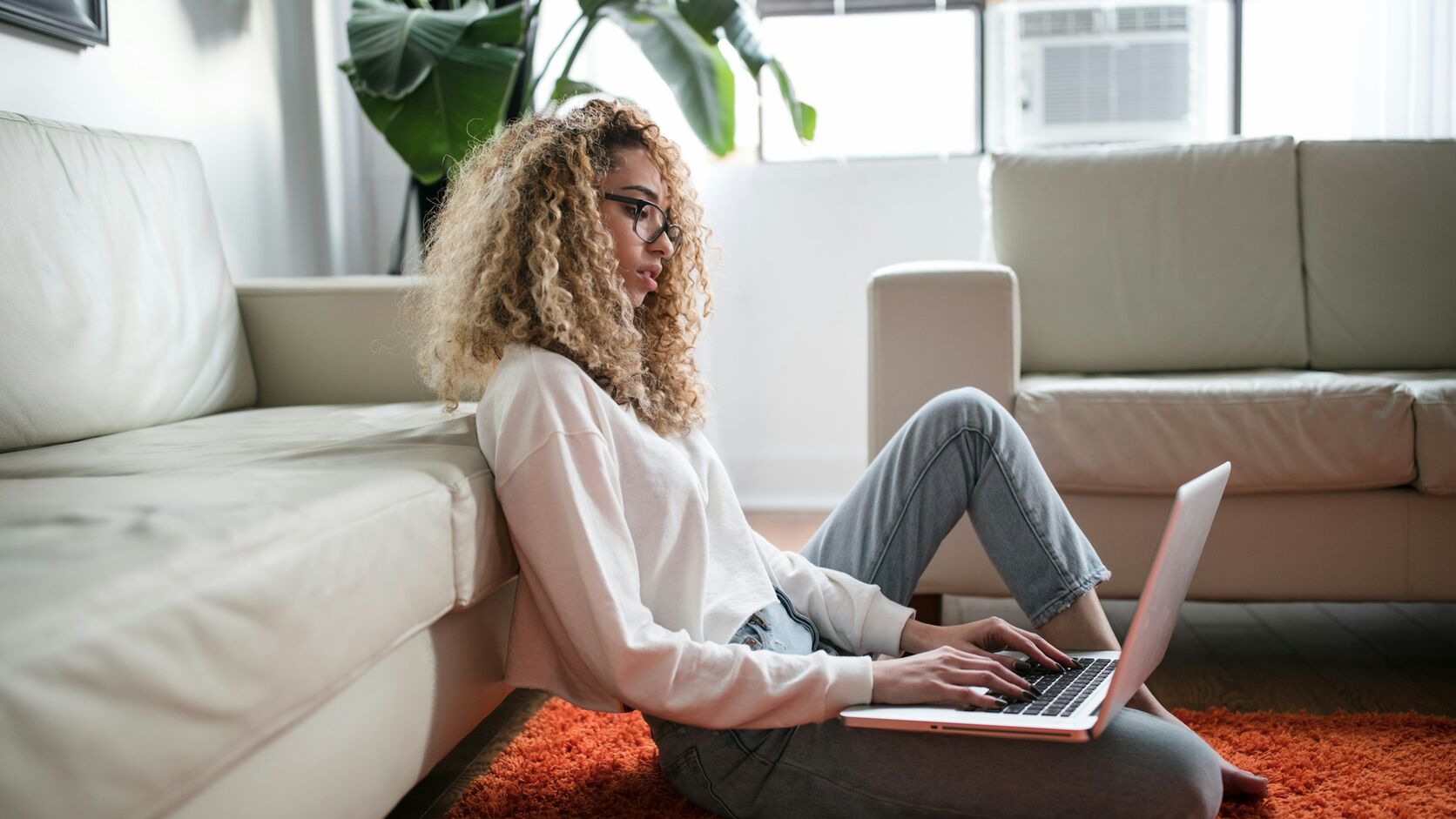 A woman with curly hair and glasses sits on the floor against a sofa, working on a laptop balanced on her lap in a sunlit living room.