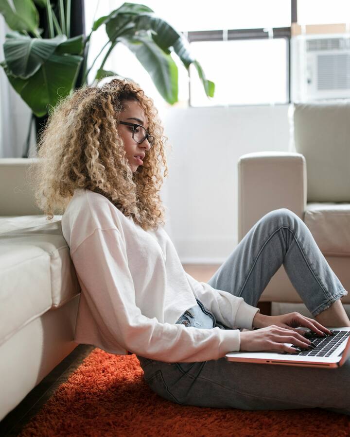 A woman with curly hair and glasses sits on the floor against a sofa, working on a laptop balanced on her lap in a sunlit living room.