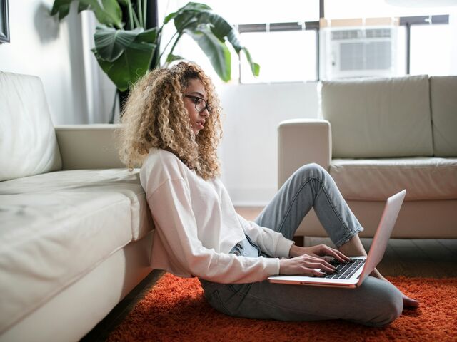 A woman with curly hair and glasses sits on the floor against a sofa, working on a laptop balanced on her lap in a sunlit living room.
