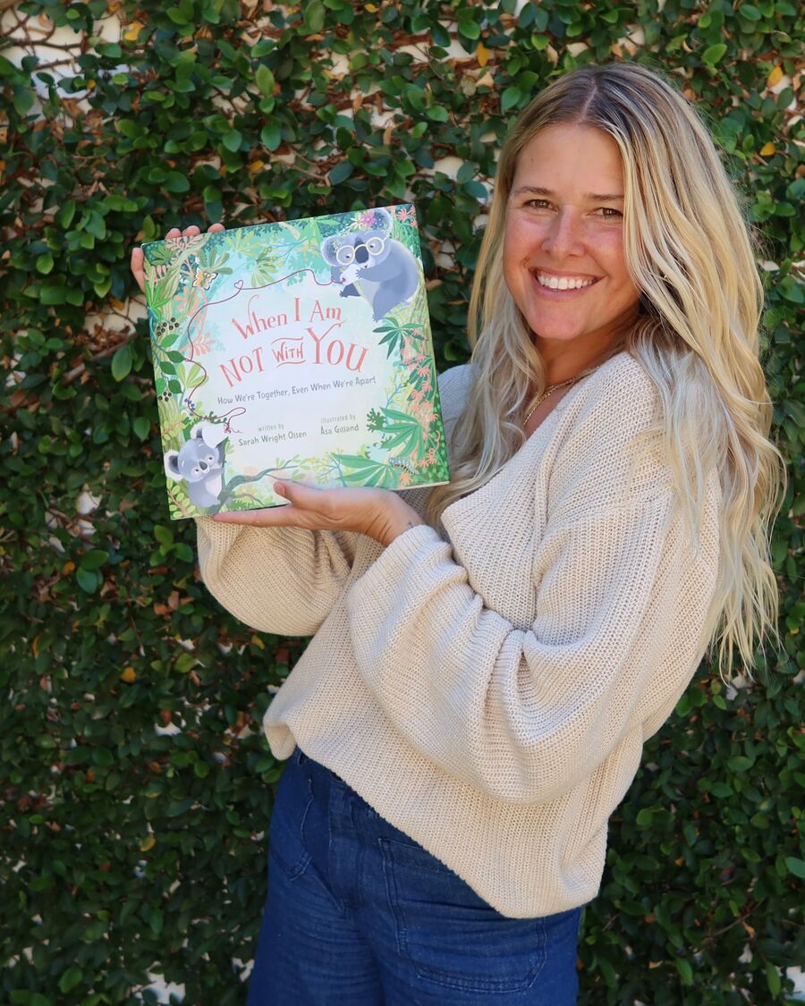 Sarah Wright Olsen smiles in front of a green hedge, holding up her children&rsquo;s book When I Am Not With You.