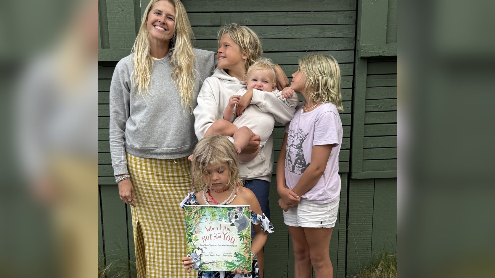 Sarah Wright Olsen stands with her four children in front of a green wooden door, her son holding the baby beside her and her youngest daughter in front holding up a copy of When I Am Not With You.