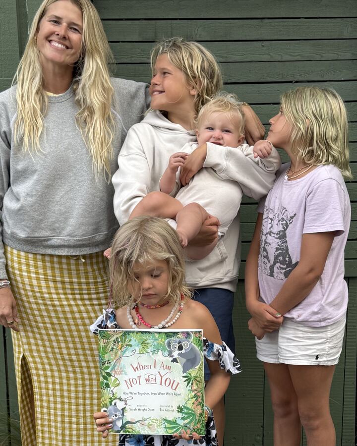 Sarah Wright Olsen stands with her four children in front of a green wooden door, her son holding the baby beside her and her youngest daughter in front holding up a copy of When I Am Not With You.