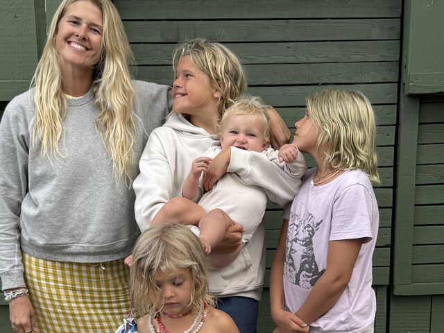 Sarah Wright Olsen smiles in front of a green hedge, holding up her children&rsquo;s book When I Am Not With You.