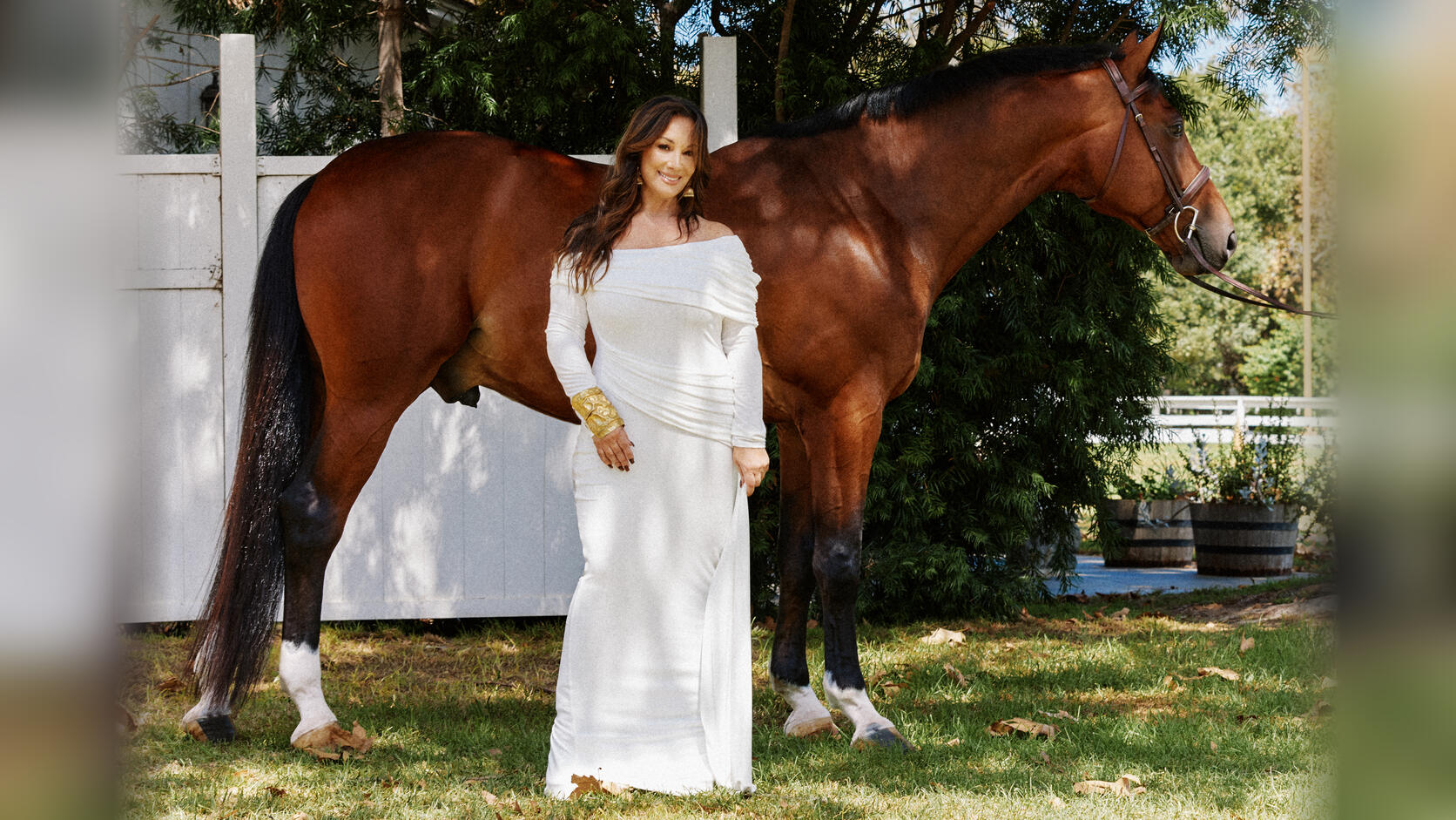 Saundra Hudson, in a white off-the-shoulder gown and a gold cuff, smiles as she stands on the grass beside a tall bay horse in a bridle, a white fence and trees behind them.