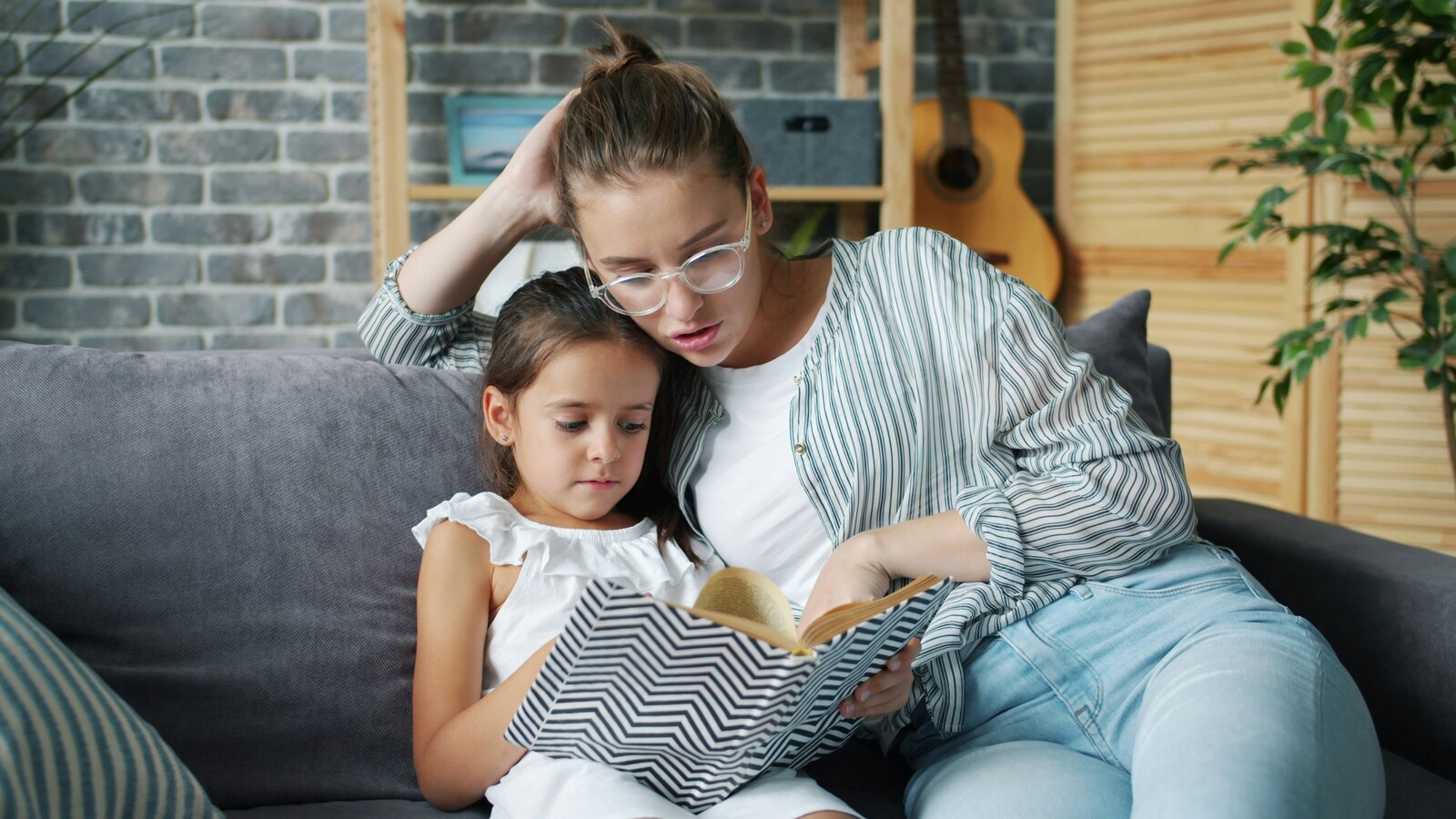 A mother in glasses and a striped shirt sits on a grey couch reading a book with her young daughter leaning against her shoulder.