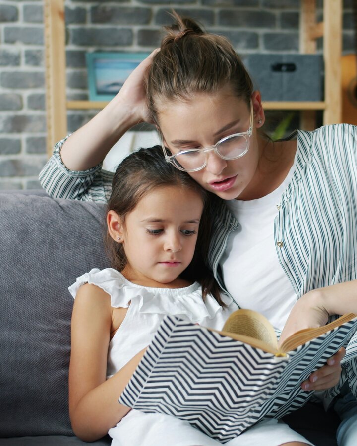 A mother in glasses and a striped shirt sits on a grey couch reading a book with her young daughter leaning against her shoulder.