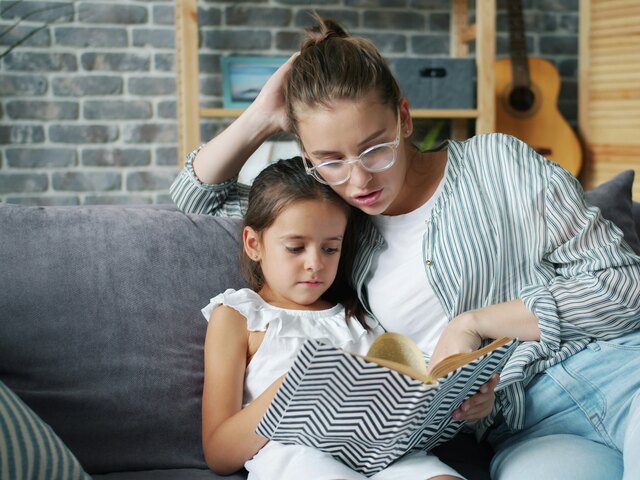 A mother in glasses and a striped shirt sits on a grey couch reading a book with her young daughter leaning against her shoulder.