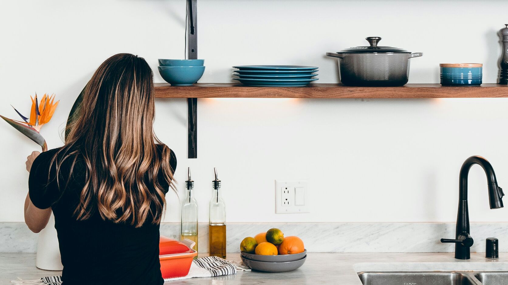 A woman with long wavy hair, seen from behind, stands at a white kitchen counter beneath a wooden shelf of blue dishes, a bowl of oranges and limes beside the sink.