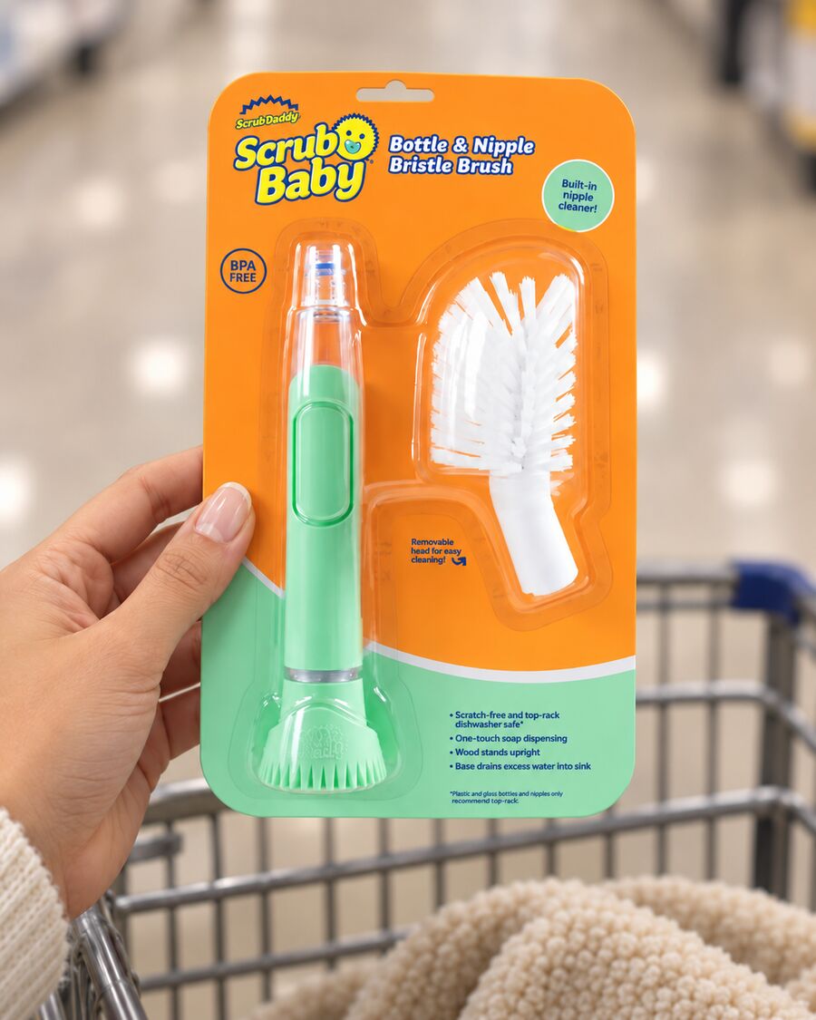 A hand holds up the Scrub Baby Bottle & Nipple Bristle Brush in its orange and mint-green package in a store aisle, a shopping cart blurred behind it.