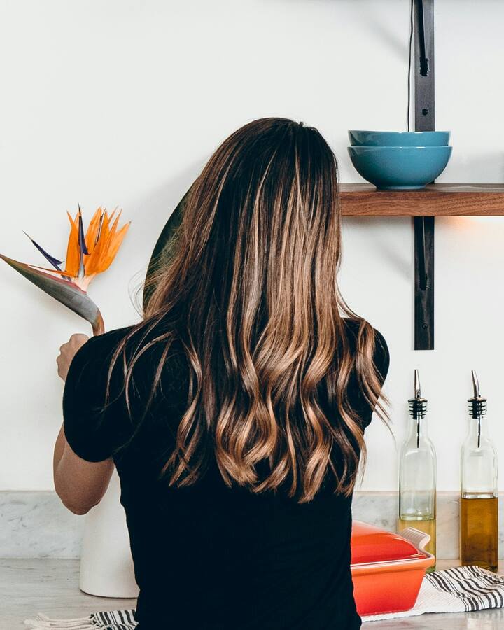 A woman with long wavy hair, seen from behind, stands at a white kitchen counter beneath a wooden shelf of blue dishes, a bowl of oranges and limes beside the sink.