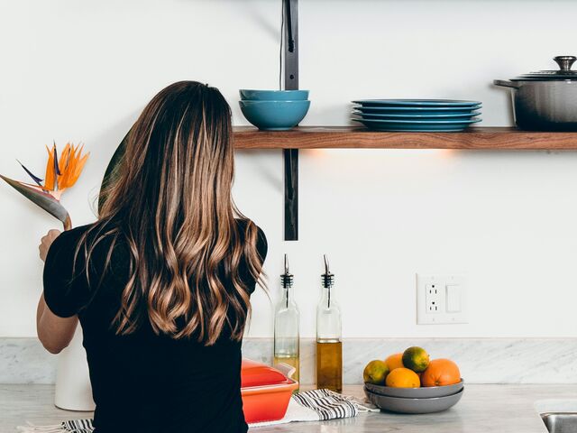 A woman with long wavy hair, seen from behind, stands at a white kitchen counter beneath a wooden shelf of blue dishes, a bowl of oranges and limes beside the sink.