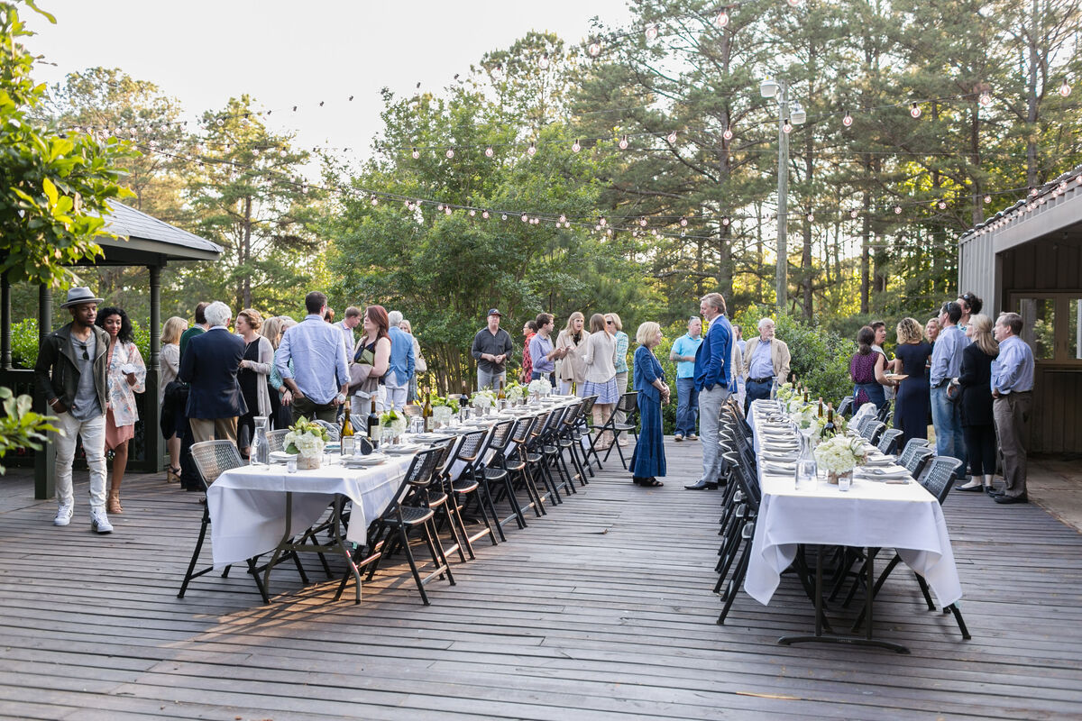 Long white-clothed tables set end to end on a wooden deck at Serenbe, guests gathered in conversation beneath strung lights and pines at golden hour.