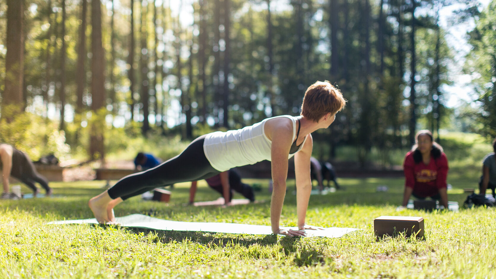 A woman holds a plank pose on a mat in a sunlit clearing at Serenbe, other students spread out on the grass behind her among tall pines.