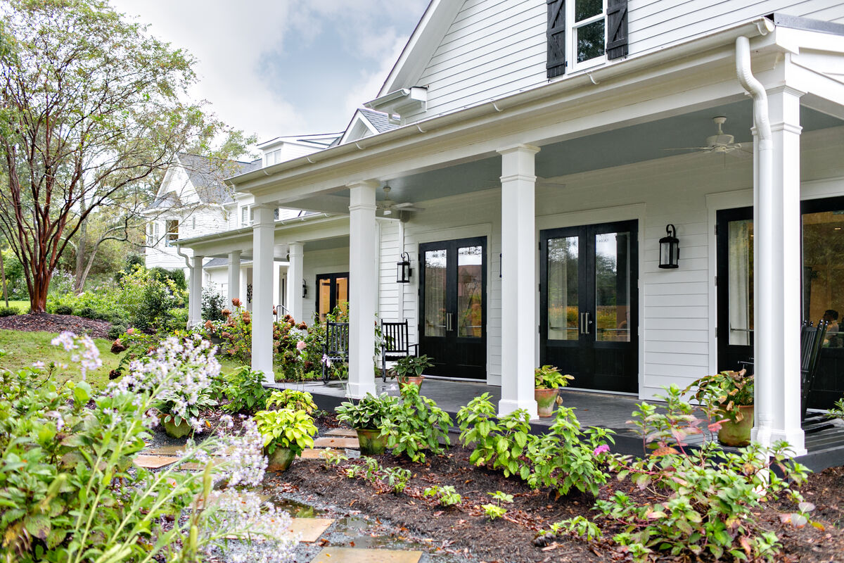 A white clapboard building at The Inn at Serenbe with a deep covered porch, black double doors, rocking chairs, and potted plants along a flagstone garden path.
