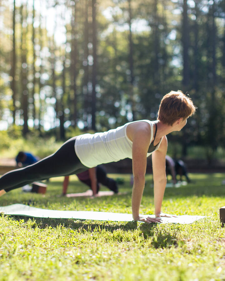 A woman holds a plank pose on a mat in a sunlit clearing at Serenbe, other students spread out on the grass behind her among tall pines.