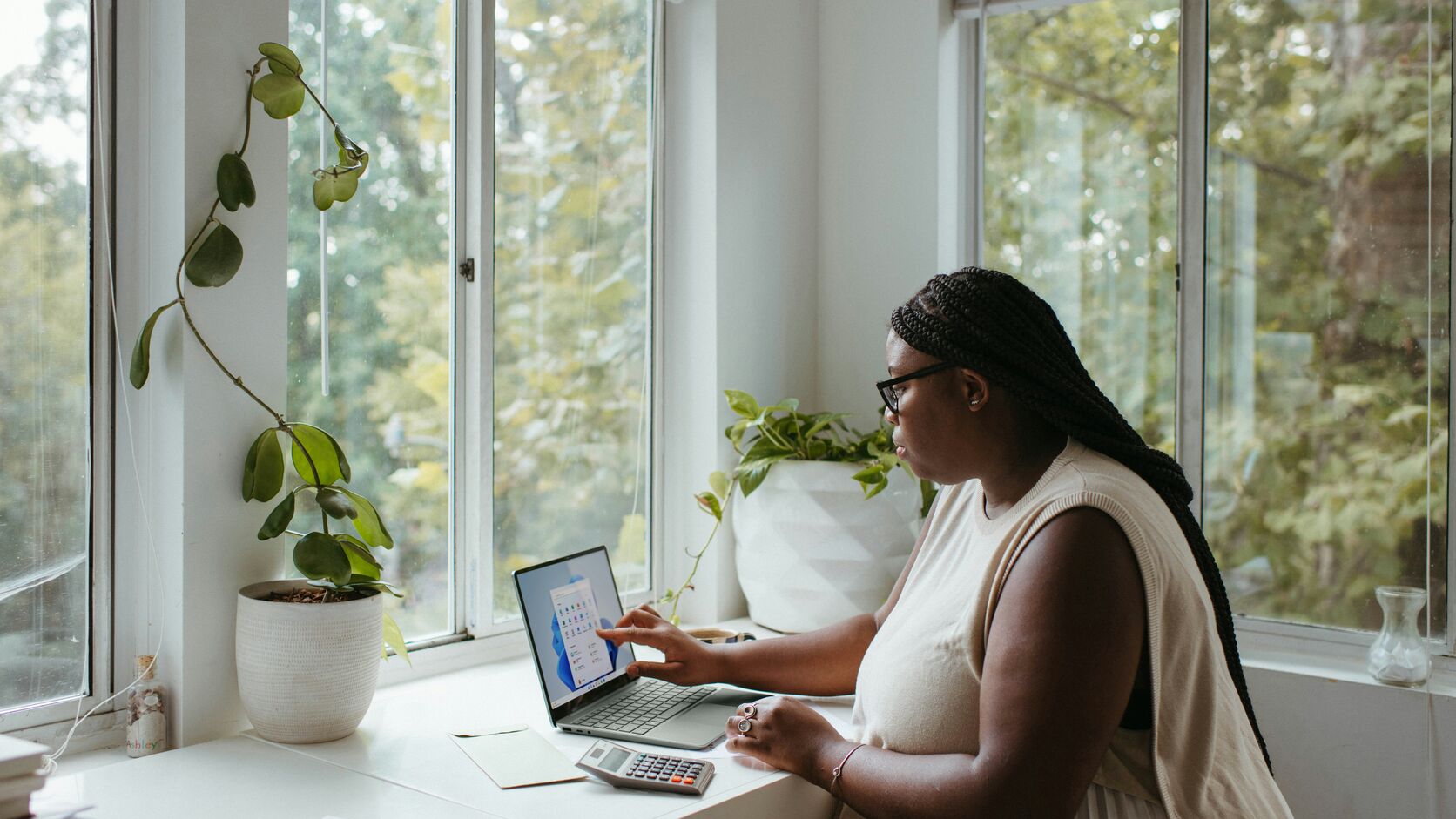 A woman with long braids and glasses works on a laptop beside a calculator at a sunlit windowsill desk surrounded by houseplants.