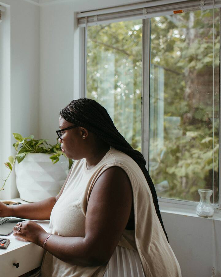 A woman with long braids and glasses works on a laptop beside a calculator at a sunlit windowsill desk surrounded by houseplants.