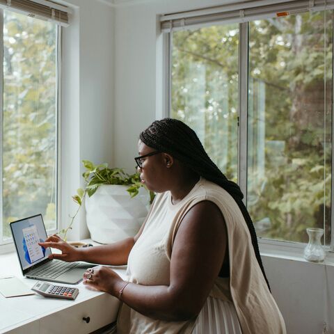 A woman with long braids and glasses works on a laptop beside a calculator at a sunlit windowsill desk surrounded by houseplants.