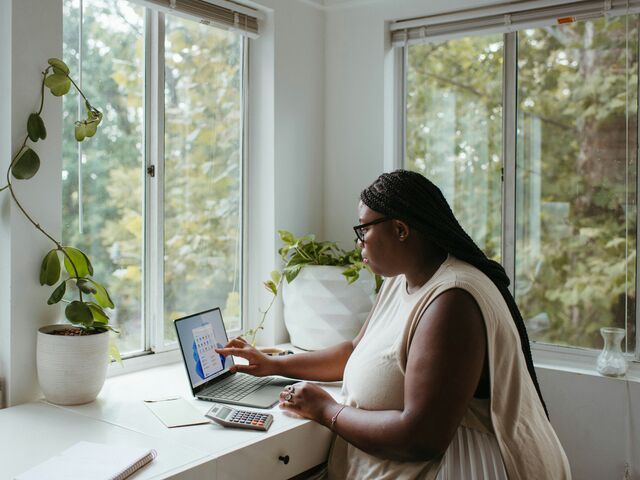 A woman with long braids and glasses works on a laptop beside a calculator at a sunlit windowsill desk surrounded by houseplants.